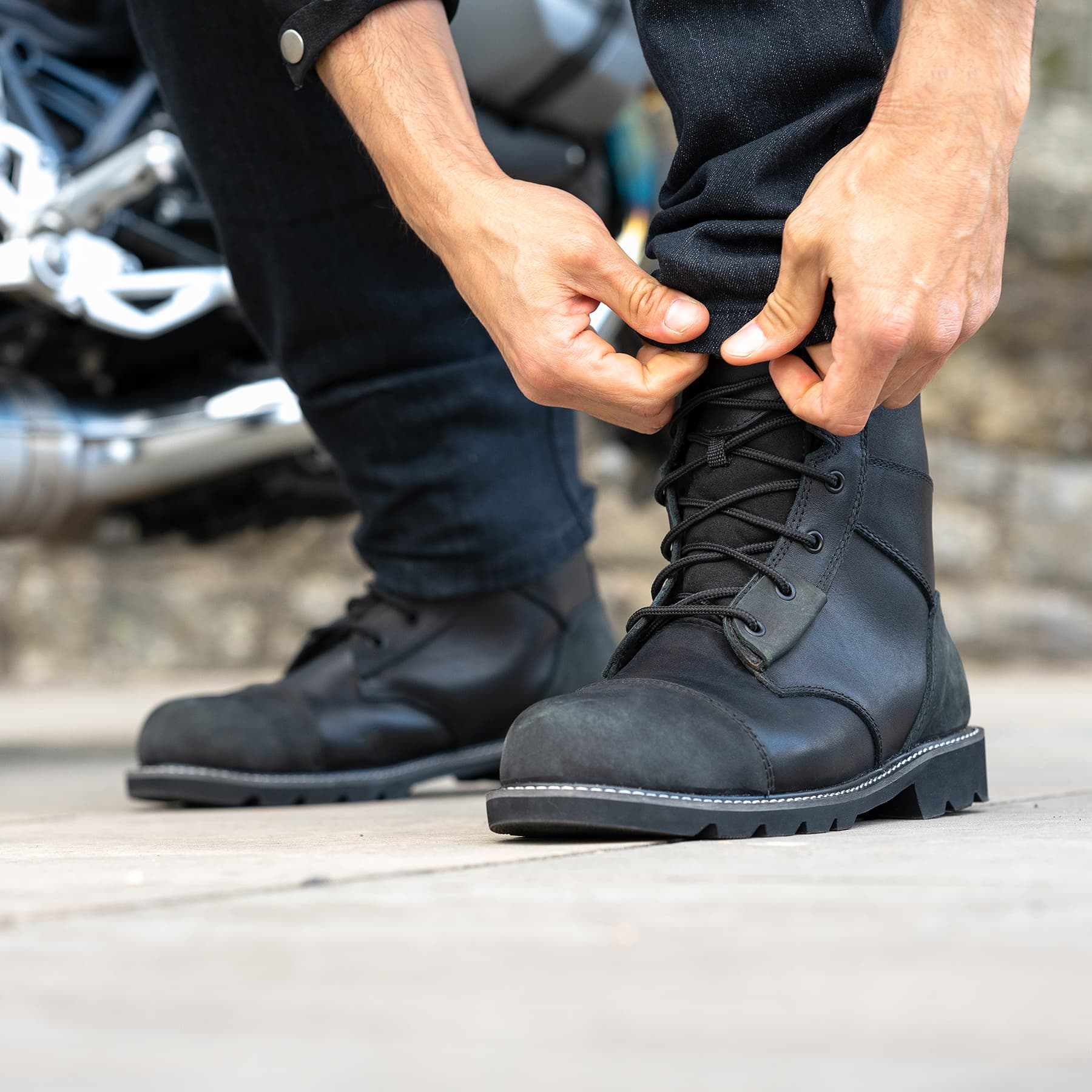 Close-up of a rider wearing vintage-style black leather motorcycle boots with reinforced toe caps and lugged soles next to a motorcycle