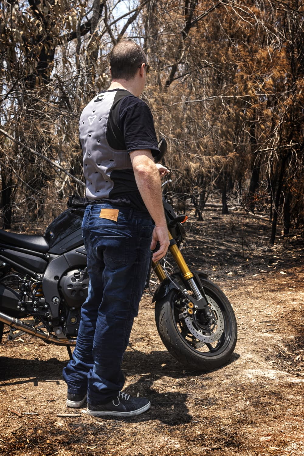 Man wearing a grey and black motorcycle protective back protector vest standing next to a black motorcycle in a wooded area