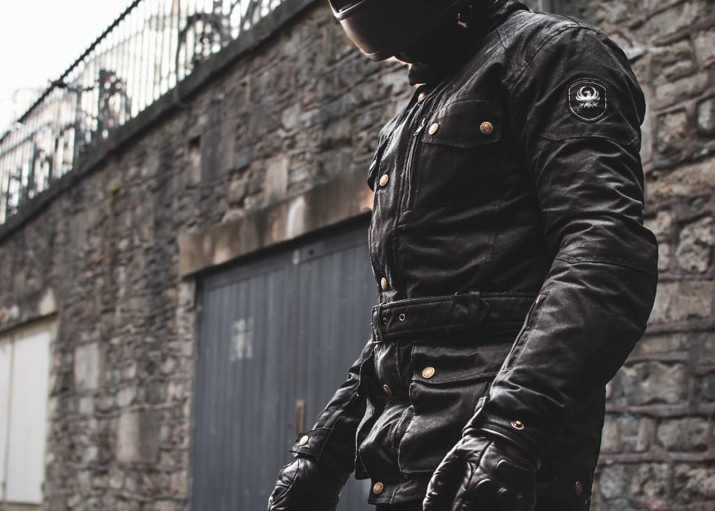 Motorcycle rider wearing a waxed leather jacket with brass buttons standing against a stone wall