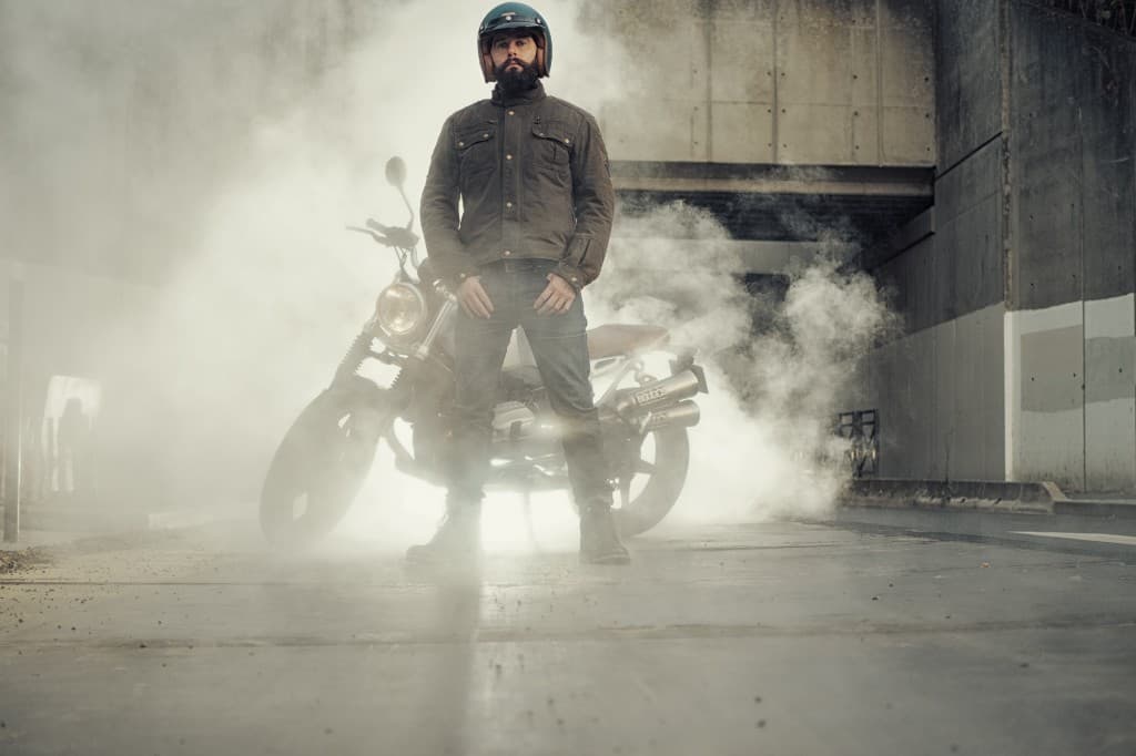 Male motorcycle rider in a brown waxed cotton winter jacket and teal open-face helmet standing beside a scrambler motorcycle surrounded by atmospheric fog in an urban underpass